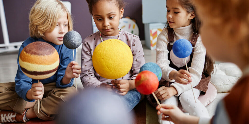 Cute little learners of primary school with models of solar system planets discussing their characteristics while studying astronomy